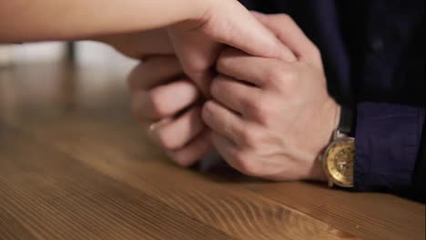 Close-Up-view-of-young-couple-holding-hands-sitting-at-the-wooden-table.-Romantic-date-of-loving-couple