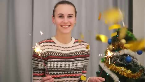 Golden-confetti-falling-on-young-smiling-woman-with-the-Christmas-tree-and-garlands-on-the-background-holding-sparklers-and