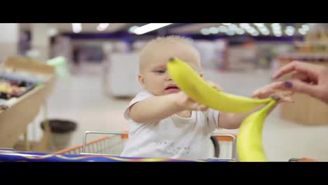 Cute-little-child-is-helping-her-mother-to-choose-bananas-in-the-supermarket-while-sitting-in-a-grocery-cart-and-holding-bananas