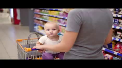 Young-mother-with-her-little-baby-sitting-in-a-grocery-cart-in-a-supermarket-is-pushing-the-cart-forward-walking-and-choosing