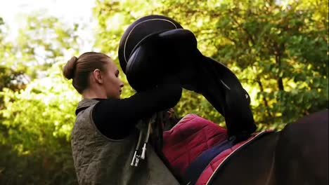 Young-beautiful-female-rider-puts-a-saddle-on-her-horse-and-prepares-animal-for-dressage