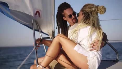 Stylish-couple-with-dreadlocks-in-white-clothes-and-sunglasses-sitting-embracing-on-the-bow-of-the-yacht-and-smiling.-Loving