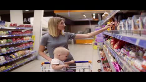 Attractive-young-mother-is-choosing-sweets-and-cakes-in-the-supermarket,-while-her-little-baby-is-sitting-in-a-grocery-cart