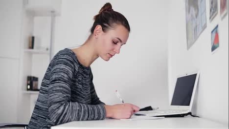 Beautiful-young-woman-sitting-by-the-table-and-writing-a-letter-using-her-pen.-Open-laptop-on-the-table.-Businesswoman-making