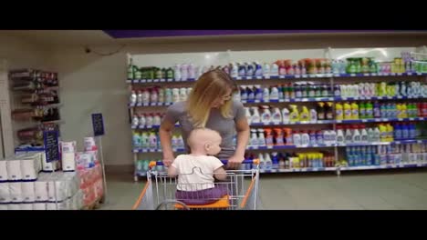 Little-baby-and-her-mother-having-fun-in-the-supermarket.-Cute-kid-is-sitting-in-a-grocery-cart,-while-her-mother-is-pushing-the