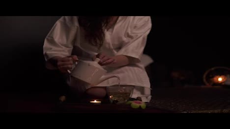 Beautiful-brunette-in-bathrobe-taking-teapot-and-pouring-tea-in-cup-drinking-herbal-tea-at-the-spa-in-slow-motion.-Thai-tea