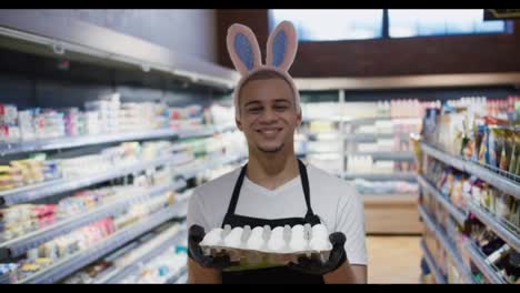 Cheerful-young-salesman-with-smile-wearing-bunny-ears-holding-eggs,-Easter-concept