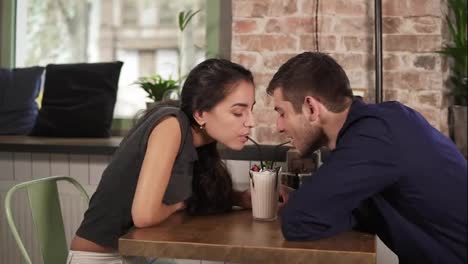 Attractive-young-couple-in-cafe-sitting-at-the-wooden-table-and-drinking-milkshake-using-straws.-Happy-couple-during-holidays