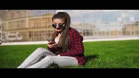 Young-woman-with-dreads-relaxing-in-park-sitting-on-the-grass-and-listening-to-music.-Hipster-girl-in-sunglasses-moving-her-body