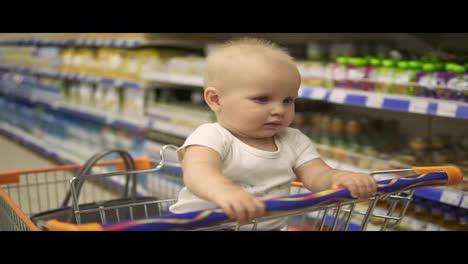 Little-baby-sitting-in-a-grocery-cart-in-a-supermarket,-while-her-mother-is-choosing-products.-Family-shopping-with-a-little