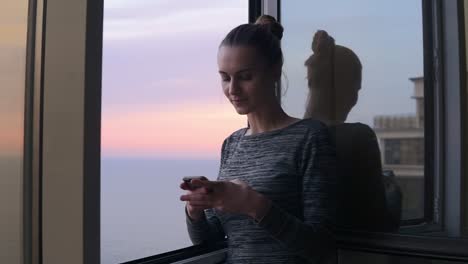 Beautiful-young-woman-sitting-at-home-by-the-open-window-with-a-smile-looking-at-phone-and-typing-a-message-during-the-sunset
