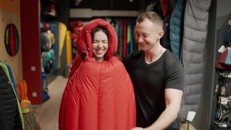A-smiling-couple-purchase-in-sportswear-store,-trying-sleeping-bag-and-make-selfie