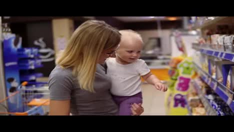 Young-mother-in-glasses-holding-her-child-in-her-arms-while-choosing-cookies-on-the-shelves-in-the-supermarket.-Thoughtful-mom