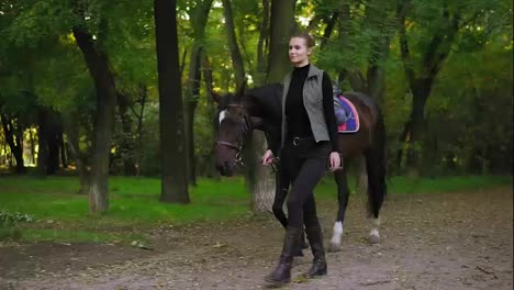 Young-happy-female-horse-rider-is-walking-with-brown-horse-with-white-spot-on-forehead-in-park-during-sunny-day-holding-leather