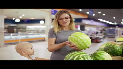 Young-mother-is-choosing-a-watermelon,-showing-it-to-her-little-child-which-is-sitting-in-a-grocery-cart.-Family-shopping-in
