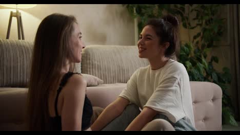 Two-young-women-having-good-time-together-in-apartment-near-sofa,-talking-and-hugging