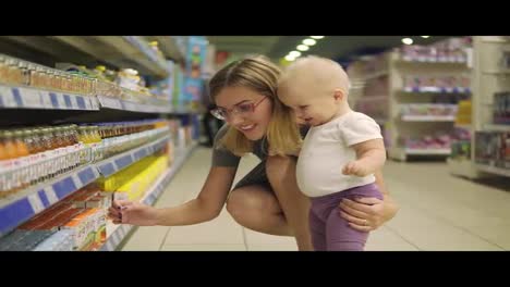 Young-attractive-mother-in-glasses-choosing-baby-food-on-the-shelves-in-the-supermarket-together-with-her-cute-child.-Mom-and