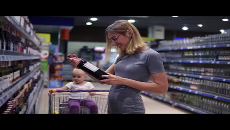 Attractive-woman-in-glasses-is-choosing-a-bottle-of-wine-in-beverages-department-in-the-supermarket,-while-her-little-baby-is