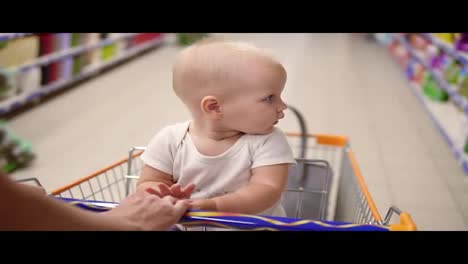 Little-cute-baby-sitting-in-a-grocery-cart-in-a-supermarket-looking-around.-The-mother's-hands-pushing-the-cart-forward-among