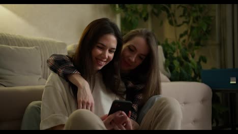 Ein-Lächelndes-LGBT-Mädchenpaar-Benutzt-Ein-Smartphone,-Während-Es-Zusammen-Auf-Dem-Boden-Sitzt