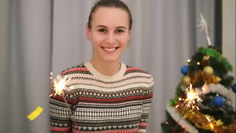 Young-smiling-woman-with-the-Christmas-tree-and-garlands-on-the-background-holding-sparklers-and-looking-to-the-camera.-Golden