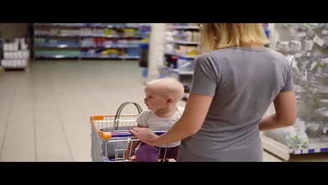 Young-mother-with-her-little-baby-sitting-in-a-grocery-cart-in-a-supermarket-is-pushing-the-cart-forward-and-choosing-products