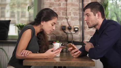 Attractive-young-couple-in-cafe-using-their-phones-sitting-at-the-wooden-table-and-drinking-milkshake-using-straws.-Happy-couple
