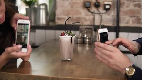 Young-man-and-woman-taking-picture-of-a-glass-with-milk-shake-with-cherry-on-the-top-and-straw-on-a-wooden-table-in-cafe