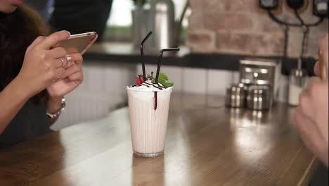 Hands-of-man-and-woman-taking-pictures-of-a-glass-with-milk-shake-with-cherry-on-the-top-and-straw-on-a-wooden-table-in-cafe