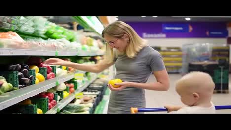 Young-mother-is-choosing-yellow-tomatoes-and-other-vegetables,-while-her-little-child-is-sitting-in-a-grocery-cart.-Family