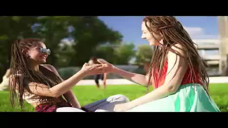 Happy-women-with-dreads-sitting-on-grass-in-summer-park.-Young-friends-talking-and-playing-outdoors-having-fun.-Mulatto-woman