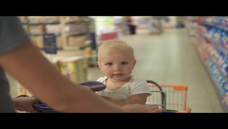 Little-baby-sitting-in-a-grocery-cart-in-a-supermarket-holding-a-toy,-while-her-mother-is-pushing-the-cart-walking-among
