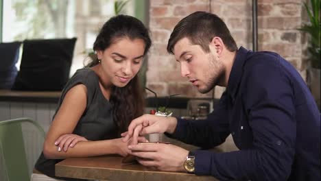 Cute-young-couple-holding-hands-and-using-phone-sitting-at-the-wooden-table-in-coffee-shot.-Romantic-date