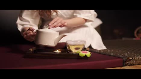 Beautiful-brunette-in-bathrobe-taking-teapot-and-pouring-tea-in-glass-cup-drinking-herbal-tea-at-the-spa-in-slow-motion.-Tea