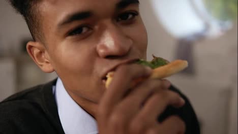 Nahaufnahme-Eines-Jungen-Afroamerikaners,-Der-Während-Des-Mittagessens-Bei-Der-Arbeit-Pizza-Genießt.-Er-Beißt-In-Ein-Stück-Und-Schaut-In-Die-Kamera