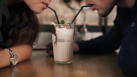 Close-Up-view-of-attractive-young-couple-in-cafe-sitting-at-the-wooden-table-and-sharing-milkshake-drinking-it-together-using-two