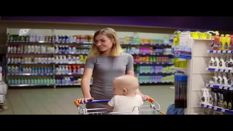 Young-mother-with-her-little-baby-sitting-in-a-grocery-cart-in-a-supermarket-is-pushing-the-cart-forward-walking-among-different