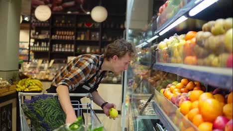 At-the-Supermarket:-Handsome-stock-clerk-wearing-black-apron,-arranging-organic-fruits-and-vegetables.-Adding-fresh-apples-on