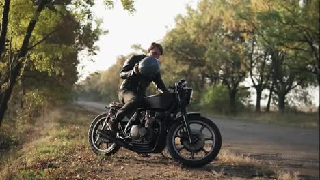 A-young-man-in-black-leather-jacket-sits-on-his-motorcycle-and-puts-a-black-helmet-to-go-on-a-journey-in-the-forest.-Slow-motion