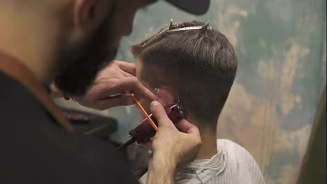 Bearded-barber-creating-a-straight-line-using-electric-trimmer-behind-a-man's-ear.-Young-handsome-caucasian-man-getting-a