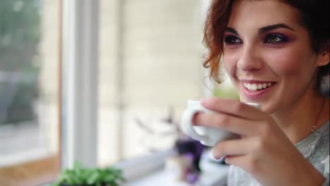 Beautiful-young-woman-enjoying-coffee-and-smiling-sitting-by-the-window-in-cafe.-Lady-with-cup-of-steaming-beverage-smiling