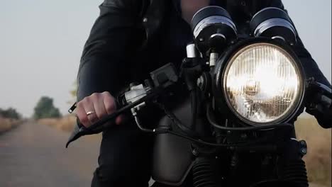 Front-view-of-a-unrecognizable-man-in-black-leather-jacket-riding-motorcycle-on-a-country-road.-Big-headlight-turned-on