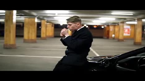 Young-man-in-a-black-suit-with-a-bow-tie-smoking-a-cigarette-sitting-at-the-bonnet-of-the-black-car-in-the-parking.-Waiting-for