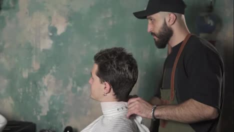 Attractive-young-man-sitting-in-a-chair-while-bearded-barber-is-preparing-him-for-a-haircut.-Stylish-barber-shot