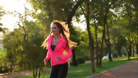Young-woman-with-long-hair-in-bright-pink-jacket-running-in-the-sunny-city-park-and-looking-in-the-camera.-Steadicam-stabilized