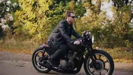 Side-view-of-a-young-man-in-sunglasses-and-leather-jacket-traveling-with-his-bike-on-and-asphalt-road-on-a-sunny-day-in-forest-in