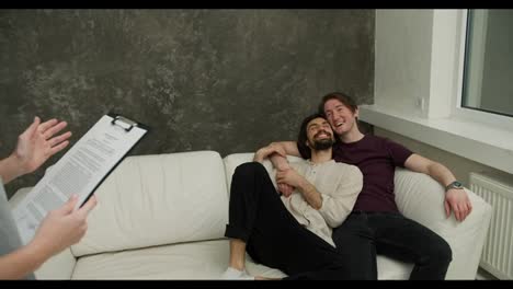 Happy-gay-couple-with-real-estate-agent-relaxing-on-sofa,-excited-about-their-new-apartment