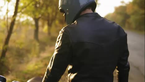Back-view-of-a-man-in-helmet-and-leather-jacket-coming-up-to-his-bike-and-starting-the-engine-while-standing-on-the-roadside-in