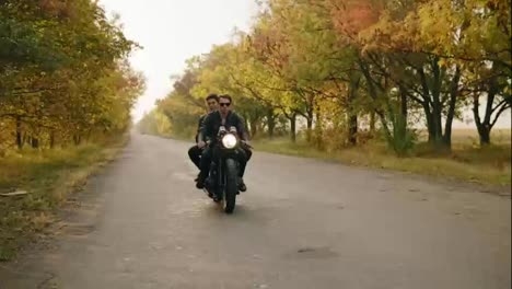 Handsome-man-in-sunglasses-riding-a-motorcycle-with-his-girlfriend-behind,-traveling-together-on-the-asphalt-road-in-forest-in