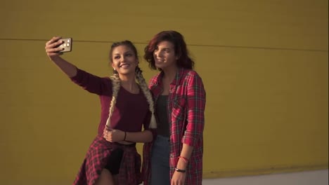 Two-best-friends-girls-having-fun-and-making-selfie-standing-by-the-yellow-wall.-Two-hipster-girls-taking-selfie-photos-with